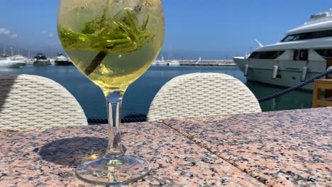 Fancy-cocktail-with-champagne-and-rosemary-by-the-sea-in-Puerto-Banus-with-yachts,-luxury-holiday-destination,-drink-on-a-tropical-vacation-with-sea-view-in-Marbella-Spain,-4K-tilting-up