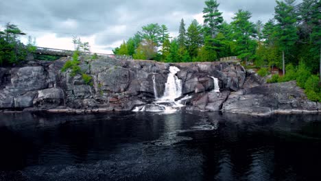 Rushing-waterfall-cascades-through-natural-bedrock-cliff-in-Northern-Ontario-with-forest-and-cloudy-sky