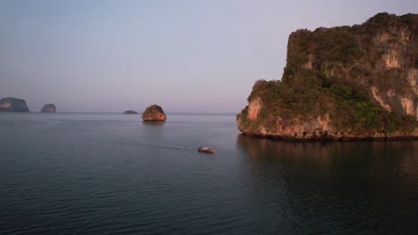wide-aerial-shot-of-a-single-thai-longtail-boat-motoring-near-large-limestome-mountains-of-Railay-Beach-in-Krabi-Thailand-during-sunrise-in-the-Andaman-Sea