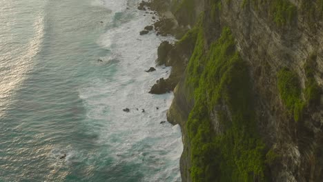 Rock-cliffs-with-large-swell-waves-crashing-onto-shore-during-sunset,-Nusa-Penida,-aerial