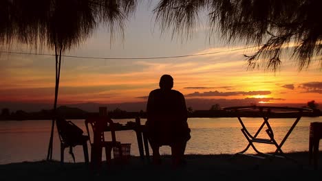 Silhouette-Eines-Mannes,-Der-Am-Flussufer-Sitzt-Und-Den-Sommersonnenuntergang-In-Kampot,-Kambodscha,-Genießt