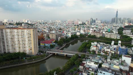 Drone-Areal-Acercándose-A-Un-Puente-Y-Canal-Que-Divide-Dos-Partes-De-La-Ciudad-En-Ho-Chi-Minh-Vietnam-Con-El-Punto-De-Referencia-81-En-La-Distancia-En-Una-Tarde-Nublada