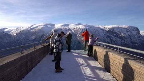 Tourists-taking-pictures-and-using-their-phones-on-tip-of-Stegastein-viewpoint-above-Aurlandsfjord-in-Norway---Walking-towards-edge-with-mountain-and-blue-sky-background