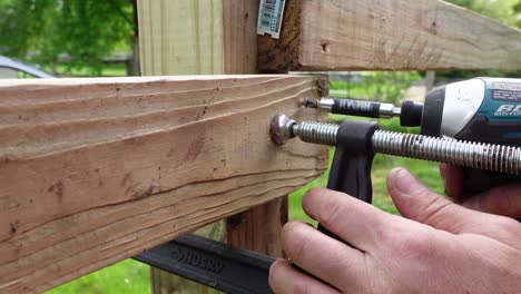 Using-metal-clamp-and-cordless-power-drill-to-build-wooden-fence,-close-up