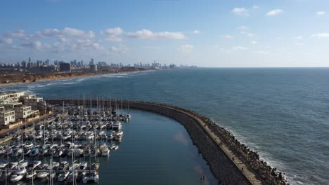 Several-tourists-walk-on-the-breakwater-that-protects-the-marina-from-the-waves-from-the-Mediterranean-Sea-with-the-skyline-of-Herzeliya-in-the-background