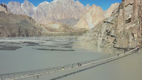 Luftdrohne-Von-Touristen,-Die-Die-Berühmte-Hussaini-Brücke-In-Hunza,-Pakistan-überqueren,-Mit-Einem-Schnellen-Fluss,-Der-Darunter-Fließt,-Und-Den-Passu-Kegelbergen-In-Der-Ferne