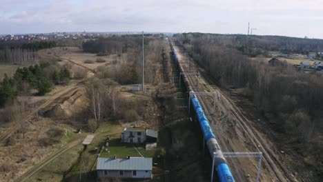 Trasportation-scene-of-industrial-train-with-many-tank-wagons-is-passing-rural-area,-leafless-trees-alongside-the-railway---aerial-drone