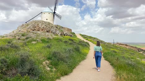 Young-blonde-country-girl-walking-towards-an-old-windmill-in-a-meadow-in-sunny-day