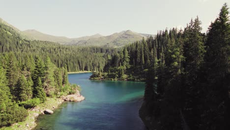 Cinematic-drone-aerial-rising-over-a-beautiful-alpine-mountain-lake-surrounded-by-woods-and-mountain-peaks