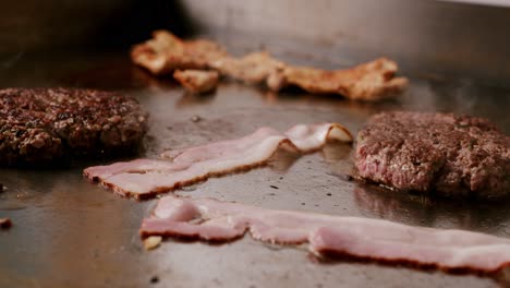 Close-up-of-uncooked-bacon-and-cooked-burger-in-hot-griddle-and-smoke-coming-out