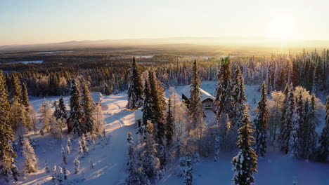 Aerial-view-across-glowing-sunrise-snow-covered-wild-rural-cabins-surrounded-by-Scandinavian-woodland