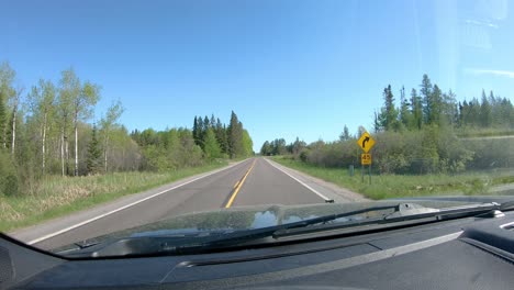 POV-while-driving-on-a-narrow-county-road-in-a-rural,-forested-area-of-northern-Minnesota-in-early-spring-on-a-bright-day