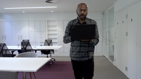 An-Asian-Indian-man-pacing-around-an-empty-office-holding-his-laptop-computer-while-talking-to-colleagues-during-an-online-conference-call-meeting