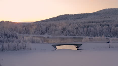 Aerial-view-flying-across-wintertime-woodland-with-vehicle-driving-along-freezing-misty-ice-lake-bridge