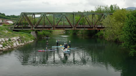 Wider-drone-shot-of-people-paddle-boarding-and-taking-a-rest-on-a-green-river-in-Slovenia-with-an-old-iron-and-rusty-train-bridge-in-the-background