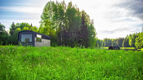 Static-shot-of-a-wooden-cottage-and-a-barrel-sauna-with-dark-clouds-passing-by-in-timelapse,-surrounded-by-green-grasslands-and-pristine-lake-at-daytime