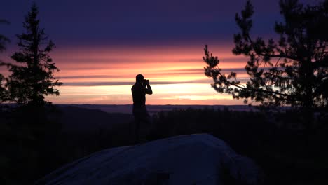 Silhouette-of-male-hiker-photographer-standing-on-a-nordic-mountain-cliff-looking-towards-horizon-with-a-summer-sunset-or-a-sunrise-over-a-big-lake-view