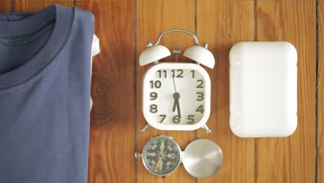 Hand-Placing-Retro-Alarm-Clock-Among-Travel-Belongings-On-Wood-Table