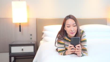 Porträt-Einer-Lächelnden,-Hübschen-Asiatischen-Frau-Mit-Sinnlichem,-Frischem,-Glücklichem-Gesicht,-Die-Auf-Ihrem-Bett-Liegt-Und-Mit-Ihrem-Smartphone-SMS-Schreibt,-Soziale-Netzwerke-Nutzt-Oder-Chattet
