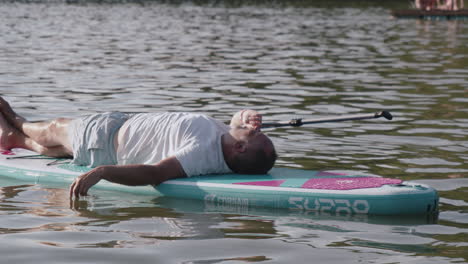 Senior-man-lying-on-paddleboard,-having-a-rest-after-paddling-on-pond-or-lake,-closeup-view