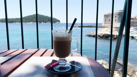 Tasse-Eisgekühlter-Latte-Mit-Blick-Auf-Das-Blaue-Meer,-Die-Felsige-Küste-Und-Die-Alten-Stadtmauern-Im-Hintergrund,-Dubrovnik,-Kroatien