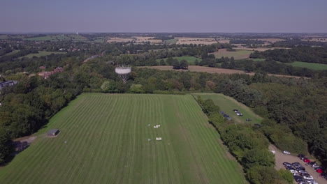 Drone-aerial-view-flying-over-freshly-cut-field-in-Trent-Park-with-car-park-and-water-tower-in-view