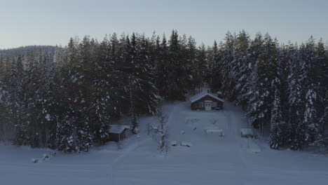 Aerial-rising-view-reveals-remote-wintertime-snow-covered-woodland-cabin-among-alpine-trees-at-sunrise