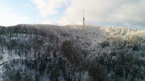 Aerial-view-of-TV-tower-on-mountain-top-surrounded-with-forest-covered-in-snow