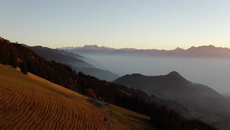 Overflying-alpine-pasture-at-sunset,-misty-atmosphere-in-the-background-with-alpine-summits-"Le-Folly",-Vaud---Switzerland