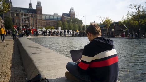 Handsome-young-man-sitting-in-front-of-Rijksmuseum-with-I-Amsterdam-sign-typing-in-a-notebook