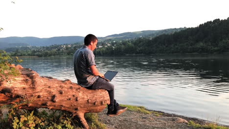 A-man-teleworks-remotely-in-nature-with-a-laptop-as-he-sits-on-a-tree-stump-looking-out-over-a-lake