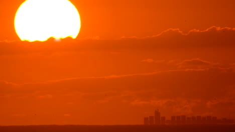 A-cargo-ship-travels-across-in-the-distance-across-the-ocean-horizon-under-a-huge-rising-sun,-glowing-the-sky-bright-orange