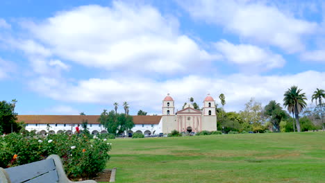 The-Santa-Barbara-Mission-building-under-blue-and-cloudy-skies-viewed-from-an-empty-bench-in-the-rose-garden-across-the-grassy-field-in-California