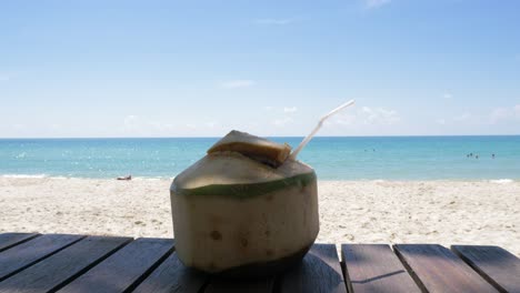Video-of-a-coconut-drink-on-a-wooden-table-on-the-beach-with-a-calm-and-sunny-ocean-view-in-the-horizon