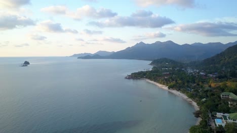 An-aerial-shot-during-a-sunset-from-a-drone-over-an-island-coast-line,-in-the-distance-there-are-visual-mountains