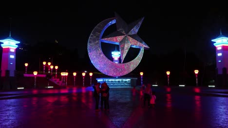 Star-and-crescent-statue-lit-up-by-colorful-lights-as-tourists-take-pictures-beneath-it-on-a-plaza-at-night