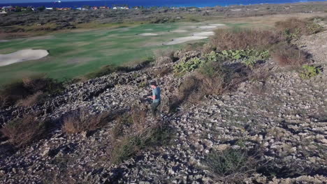 A-nature-photographer-stepping-over-a-large-crack-in-the-volcanic-rock,-overlooking-a-golf-course-in-Aruba