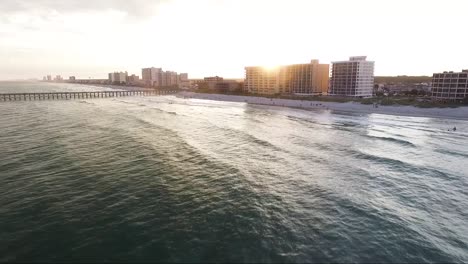 Drone-flying-towards-beach-at-sunset-near-a-pier