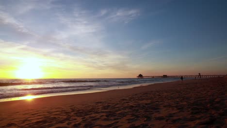 Ein-Wunderschöner-Zeitraffer-Des-Strandes-Während-Eines-Gelb-blauen-Sonnenuntergangs-Mit-Dem-Huntington-Beach-Pier-Im-Hintergrund-In-Surf-City,-USA,-Kalifornien