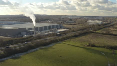 Rising-aerial-drone-shot-of-factory-with-smoking-chimney-over-fields-and-motorway