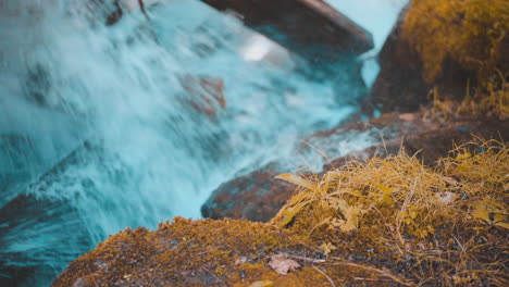 Rushing-Water-Rapids-Crashing-Into-Large-Rocks-in-Background-with-Moss-Covered-Rocks-in-Foreground-During-Autumn-4K-ProRes