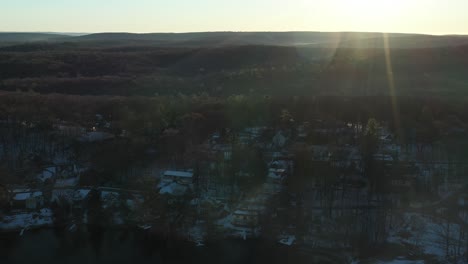 Drohne-Zieht-Bei-Schneebedecktem-Sonnenaufgang-Im-Winter-über-Ländliche-Häuser-Im-Wald-Am-Ufer-Eines-Sees-Hoch