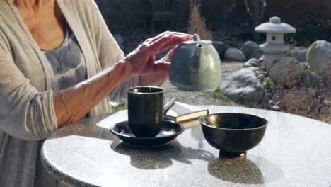 A-woman-placing-an-herbal-tea-bag-into-a-cup-and-pouring-hot-water-from-a-teapot-then-letting-it-steep
