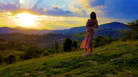 Sunset-over-the-mountains-and-a-cloudy-sky-with-a-girl-posing,-spinning-and-jumping-on-a-grassy-hill