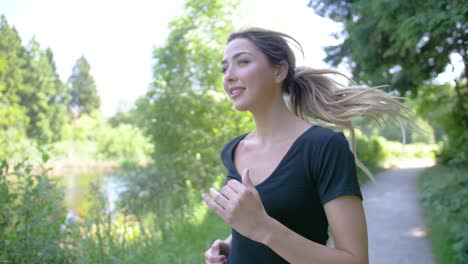 A-happy-young-woman-enjoys-the-scenery-of-a-park-setting-as-the-runs-along-a-path-near-a-pond-on-a-beautiful-sunny-day