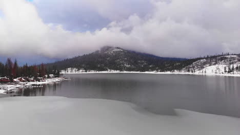 Luftrückzug-über-Einem-Teilweise-Zugefrorenen-See-Und-Schneebedeckten-Bewaldeten-Ufern-Mit-Einem-Wunderschönen-Schneebedeckten-Berg,-Der-Von-Wolken-Am-Blauen-Himmel-Im-Hintergrund-Berührt-Wird