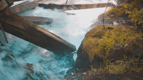 Rushing-Water-Rapids-Crashing-Into-Large-Damaged-Wooden-Beams-with-Moss-Covered-Rocks-During-Autumn-4K-ProRes