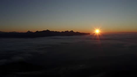 Flying-high-and-fast-in-reverse-over-sea-of-clouds-at-sunset,-the-Alps-in-the-background---Switzerland