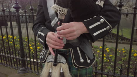 Close-up-of-a-male-Scottish-bagpipers-hands-as-he-plays-the-bagpipes-next-to-a-black,-ornate-metal-fence-with-Princes-Street-gardens-in-the-background,-Edinburgh,-Scotland