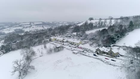 Aerial-tracking-towards-a-quaint-English-village-with-colourful-houses-and-thatched-cottages-all-covered-in-a-blanket-of-winter-snow